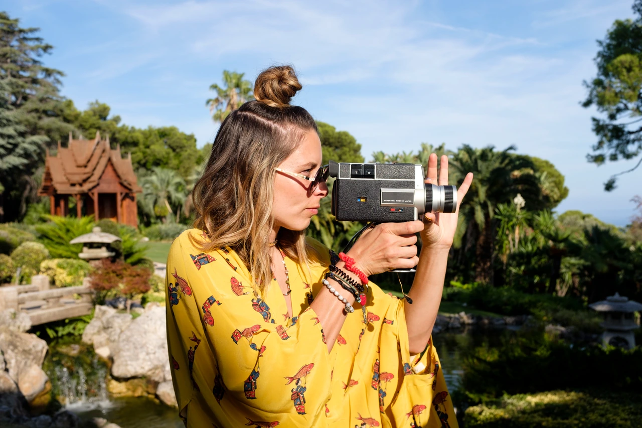 A person in yellow clothing looks through a vintage movie camera in a garden, from the Blondish series.